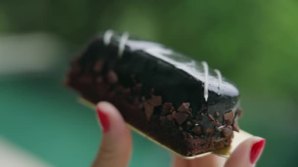 Primer plano de una chica con una manicura de color rojo brillante sosteniendo una apetecible eclair oscuro cubierto con virutas de chocolate y café sobre el fondo de hermosos árboles verdes en desenfoque. Movimiento lento. — Vídeos de Stock