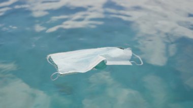 Tıbbi maske deniz yeşili sularda yüzüyor ve güneş parlıyor. Koronavirüs salgınının sonuçları çevreyle ve tek kullanımlık maskeler yüzünden oluşan çöplerle ilgilidir..