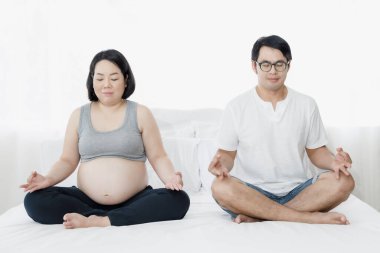 Koca ve hamile kadın beyaz yatak odasında yoga yapıyorlar ve arka planda doğal ışık pencereleri var.  