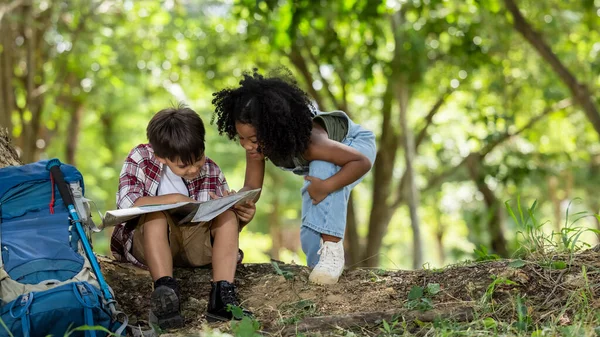 African American ethnicity boy and girl children wearing plaid shirt and backpack sitting at tree base talking and looking down on the map travel on hand in the park background hiking adventure summer holiday concept