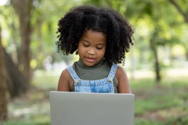 children girl African American ethnicity black skin sitting on tree base use Laptop computer to chat with friends and via video call with 5G internet signal in the park, social distance concept
