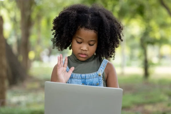children girl African American ethnicity black skin sitting on tree base use Laptop computer via video call and holding hands goodbye friends and she looks sad with 5G internet signal in the park, social distance concept