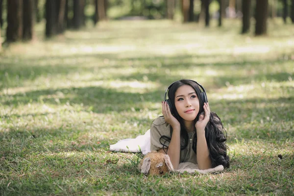 美丽的亚洲女人躺在松林花园里放松地听着耳机蓝牙的音乐 还有浅橙色的工艺背景 孤独的大自然人的概念 — 图库照片