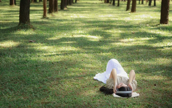 美丽的亚洲女人躺在松林花园里放松地听着耳机蓝牙的音乐 还有浅橙色的工艺背景 孤独的大自然人的概念 — 图库照片