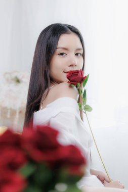 Güzel bir Asyalı kadın elinde tek bir kırmızı gül tutarken nazikçe gülümsüyor. Beyaz bir elbiseyle önplanda bulanık çiçekler olan parlak bir arkaplanda romantik bir sevgililer günü konsepti için poz veriyor..