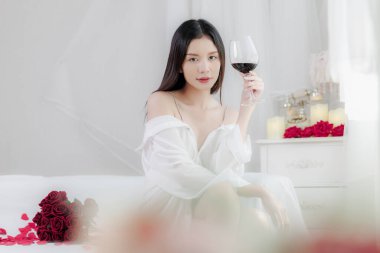 Beyaz gömlekli çekici Asyalı kadın gül yapraklarıyla süslenmiş bir yatakta bir bardak kırmızı şaraptan hoşlanıyor. Romantik ortamda sevgililer günü kutlaması için mumlar ve çiçekler olacak..
