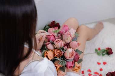 Yüksek açılı yakın çekim, bir kadının ellerinde bir buket eski renkli gül (tuğla turuncusu, tozlu pembe) üzerine odaklanıyor. Bacaklarını ve tenini gösteriyor. Estetik ve sanatsal ruh hali.