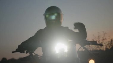 Açık havada park etmiş, arka planda bulanık bir günbatımı olan modern bir motosikletin ışıklarına yakın çekim. Kask penceresini ayarlayan bir motosikletçi. 
