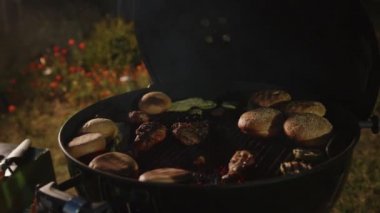 Lezzetli hamburger çörekleri ve sebzeler, arka planda bahçe çiçekleri ile ızgara yapıyorlar..