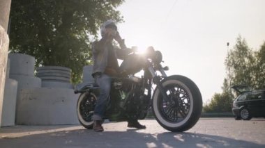 Kot pantolon giyen yakışıklı bir motosikletçi, deri ceket, özel motosikletinin üzerinde oturan şık botlar, arka planda güneş parlayan gümüş kaskını ayarlıyor..