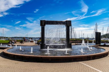 Kristiansand, Norveç-12 Ağustos 2025: Otterdalsparken, Kristiansand 'da sanatçı Kjell Nupen' in granit çeşmesi. Yat limanı, insanlar, yelkenliler ve yaz yeşiliyle çevrili..