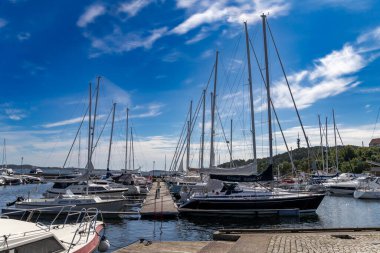 Kristiansand, Norveç-12 Ağustos 2025: Birçok yelkenli Kristiansand marinasına demirlemiş, sakin su, ahşap iskele ve limanın üzerinde dinamik bulutlar olan mavi gökyüzü.