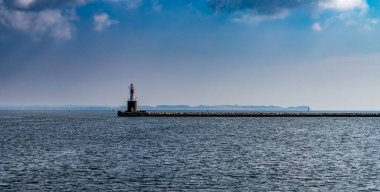 Aarhus, Danimarka-15 Ağustos 2025: Durgun denizde iskelede duran kırmızı ve beyaz deniz feneri, uzak yelkenli ve arka planda kıyı ufkuyla dalgakıran.