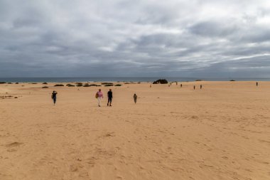 İspanya, Kanarya Adaları, Fuerteventura-5 Şubat 2026: Turistler kıyı manzarasında bulutlu gökyüzü altında geniş kum tepelerini aşarak okyanus ufkuna doğru yürürler.