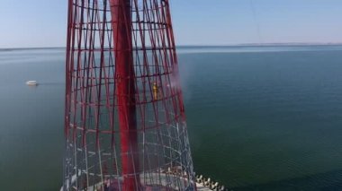 Yazın açık denizde sıcak spreyle temizleyen işçiyle birlikte yükseltilmiş fener havası. Metalik ızgaralardan yapılmış gökyüzü gözlüklü fenere çarpıyor. Üniformalı işçiler güneşli bir günde sıcak suyla tırmanıp temizliyor. 