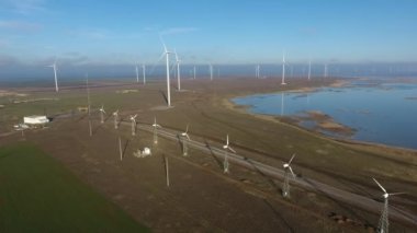 Yazın yüksek rüzgar enerjisi direklerinden oluşan hava şeritleri ve dönen vanalar. Yüksek beyaz sütunların çizgileri, dönen bıçaklarla yazın güneşli bir günde rüzgâr enerjisi topluyor. Deniz güzel görünüyor..