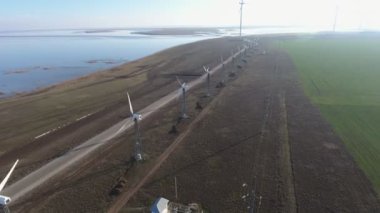 Yazın dönen bıçaklarla rüzgar enerjisi direklerinin hava görüntüsü. Güneşli bir yaz gününde deniz kenarında rüzgar enerjisi toplayan bir sıra yüksek beyaz direğin kuş bakışı görüntüsü. Deniz kenarı iyi görünüyor..