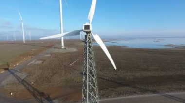 Yazın, rüzgâr enerjisi direkleri ve dönen bıçaklarla dolu bir sıranın havadan çekilmiş görüntüsü. Güneşli bir yaz gününde deniz kenarında rüzgâr enerjisi toplayan yüksek beyaz direklerin oluşturduğu bir sıra hayret verici kuş bakışı. Gökyüzü mavi.
