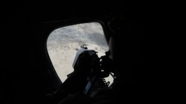Asker makineli tüfeği tutuyor ve uçan bir helikopterin kapağına bakıyor. Kasklı, modern makineli tüfekli ve güneşli bir yaz gününde askeri helikopterin kapağına bakan askerin etkileyici görüntüsü. Yeşil alan görüldü.