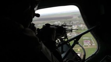 Yazın elinde makineli tüfekle uçan bir helikopterin kapağına bakan asker miğferli asker, günümüzün makineli tüfeğini tutuyor ve yazın güneşli bir günde askeri helikopterin kapağına bakıyor. Küçük evler görünüyor.