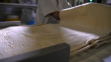Büyük hamur tabakası, modern bir fırın fabrikasında üniformalı bir işçi tarafından açılır. Lastik eldivenli ve üniformalı bir işçi tarafından açılmış geniş hamurdan yapılmış fevkalade bir yakın plan. Bir fırın fabrikasında reçelli kruvasan pişirmek için..