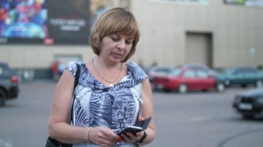 İlkbaharın sonlarında güzel bir sarışın kadın, park yerinde telefon ekranına dokunuyor. Hafif elbiseli, meşgul bir sarışının baharda büyük bir otoparkta akıllı telefon ekranına dokunuşunu hayret verici bir şekilde görüyor. Orada süpermarketler ve otomobiller görülüyor..