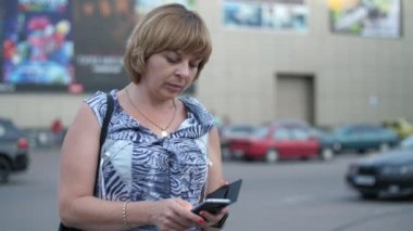 Sakin sarışın kadın yavaş çekimde park yerinde telefon ekranına dokunuyor hafif elbiseli hoş sarışın bir kadının ağır çekimde büyük bir otoparkta akıllı telefon ekranına dokunuşunun neşeli görüntüsü. Orada binalar ve otomobiller görülüyor..