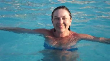 Bikinili mutlu sarışın bir kadın ağır çekimde havuzda duruyor ve dinleniyor mavi bikinili mutlu sarışın bir kadının açık mavi sularda yüzen bir havuzun basamaklarında durup gülümsemesi ve ağır çekimde hayatın tadını çıkarması.. 