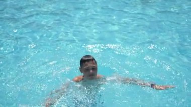 Neşeli çocuk, gülümseyen ve yüzme havuzunda sıçrayan sersemletici görüntüsüyle harika bir esmer çocuk, tayfa tıraşlı, zıplayan, el kaldıran, dalgalı bir havuzda güneşli bir günde turkuaz sularda yüzen ve gülümseyen. 