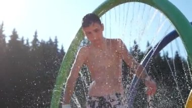 Mutlu çocuk dışarıda, dağlarda yuvarlak şekilli bir duşta dans ediyor. Yazın güneşli bir yaz gününde, dağlardan fışkıran su akıntılarıyla yeşil bir çember içinde dans eden ve gülümseyen bir çocuğun neşeli görüntüsü..