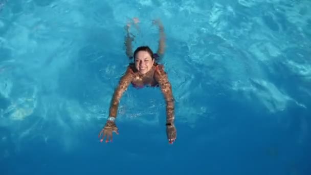 幸せな女性が笑顔と青い水でプールでの水泳のブレインストークビキニで楽観的な公正な髪の女性の陽気な景色笑顔と夏の晴れた日にライトブルーの水でプールでの水泳の平泳ぎ — ストック動画