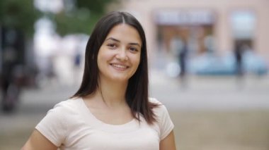 Neşeli esmer kız, yazın güneşli bir günde yeşil sokakta ayakta ve gülümsüyor. Uzun saçlı, beyaz tişörtlü güzel bir kız. Kalçaları üzerinde duruyor ve yazın sokakta mutlu bir şekilde gülümsüyor.         