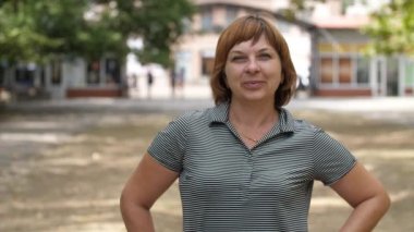 Yazın havadar ve güneşli bir parkta ayakta duran ve gülümseyen mutlu sarışın kadın gri bluzlu neşeli, açık renk saçlı, ayakta duran, gülümseyen ve ellerini yazın evi olan büyük bir parkta kalçalarında tutan bir kadın manzarası heyecan verici..