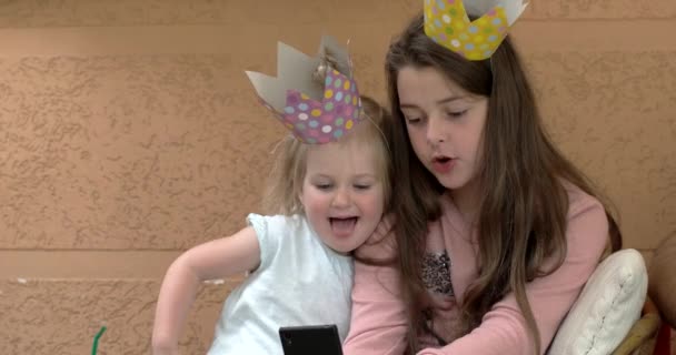 Две милые сестрички в коронах развлекаются с мобильным телефоном и делают смешное селфи — стоковое видео