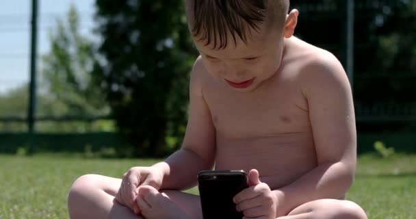 Liten pojke sitter på en grön äng med en mobiltelefon — Stockvideo