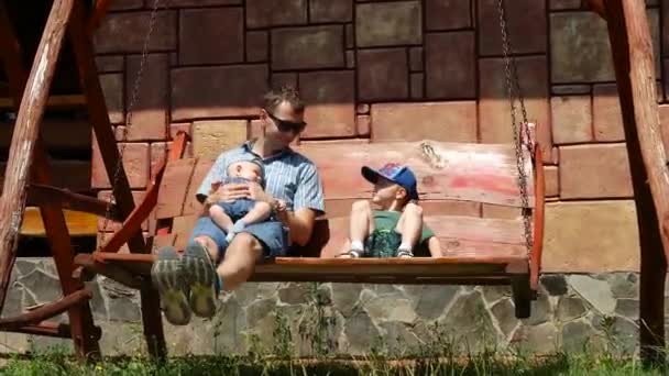 Jovem pai caucasiano com seus filhos jovens descansando em um balanço de corrente de madeira — Vídeo de Stock