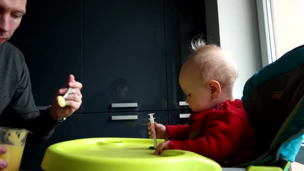 Молодий батько годує дитину сином у високому кріслі на кухні — стокове відео