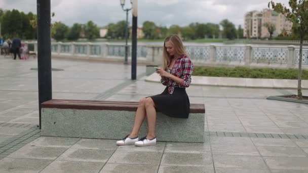 Jeune femme avec smartphone assis sur un banc dans le parc — Video