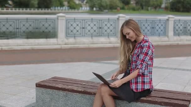 Mujer joven trabaja en la computadora portátil moderna en el parque de la ciudad sonriendo — Vídeos de Stock