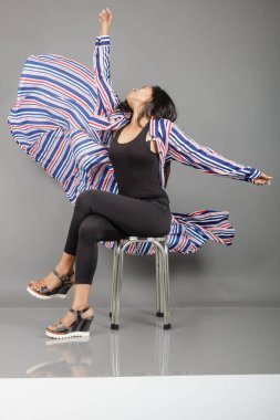 Dişi bir dansçı metal bir taburede oturur, çizgili uzun paltosu etrafında dönerken canlı ve anlamlı bir poz verir. Moda, yaşam tarzı, güçlendirme ve yaratıcı hareket kavramları için mükemmel..