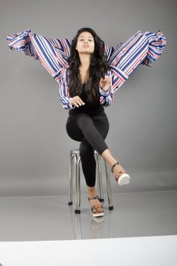 Dişi bir dansçı metal bir taburede oturur, çizgili uzun paltosu etrafında dönerken canlı ve anlamlı bir poz verir. Moda, yaşam tarzı, güçlendirme ve yaratıcı hareket kavramları için mükemmel..