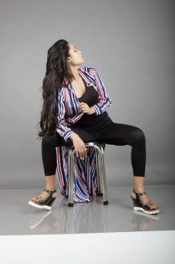 Mavi, kırmızı ve beyaz çizgili ceketli, siyah bluzlu, saçları yukarı doğru uçan, saç ürünleri ve moda reklamları için ideal bir taburede oturan kendine güvenen genç bir kadın manken..