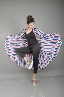 Siyah tayt, topuklu, siyah üst ve kırmızı, mavi ve beyaz çizgili tunik giyen güzel, genç bir kadın. Gülüyor, dönüyor, zıplıyor, neşeyle dans ediyor ve bir tabureyle poz veriyor.