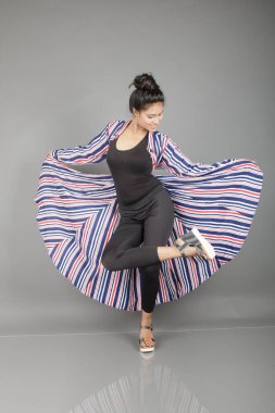 Siyah tayt, topuklu, siyah üst ve kırmızı, mavi ve beyaz çizgili tunik giyen güzel, genç bir kadın. Gülüyor, dönüyor, zıplıyor, neşeyle dans ediyor ve bir tabureyle poz veriyor.