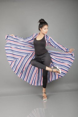 Siyah tayt, topuklu, siyah üst ve kırmızı, mavi ve beyaz çizgili tunik giyen güzel, genç bir kadın. Gülüyor, dönüyor, zıplıyor, neşeyle dans ediyor ve bir tabureyle poz veriyor.