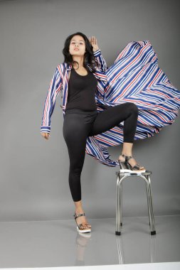 Siyah tayt, topuklu, siyah üst ve kırmızı, mavi ve beyaz çizgili tunik giyen güzel, genç bir kadın. Gülüyor, dönüyor, zıplıyor, neşeyle dans ediyor ve bir tabureyle poz veriyor.