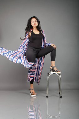 Siyah tayt, topuklu, siyah üst ve kırmızı, mavi ve beyaz çizgili tunik giyen güzel, genç bir kadın. Gülüyor, dönüyor, zıplıyor, neşeyle dans ediyor ve bir tabureyle poz veriyor.