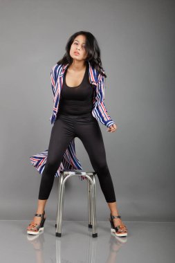 Siyah tayt, topuklu, siyah üst ve kırmızı, mavi ve beyaz çizgili tunik giyen güzel, genç bir kadın. Gülüyor, dönüyor, zıplıyor, neşeyle dans ediyor ve bir tabureyle poz veriyor.