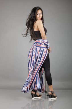 Siyah tayt, topuklu, siyah üst ve kırmızı, mavi ve beyaz çizgili tunik giyen güzel, genç bir kadın. Gülüyor, dönüyor, zıplıyor, neşeyle dans ediyor ve bir tabureyle poz veriyor.