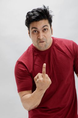 Kırmızı tişörtlü kızgın genç adam hayal kırıklığını ifade ediyor ve orta parmağını gösteriyor. Beyaz arka plandaki stüdyo portresi el hareketi kullanarak güçlü duygu ve tepki kavramını gösteriyor.
